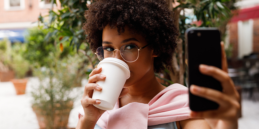 Woman holding a cup of coffee taking a selfie, trees in backgroun